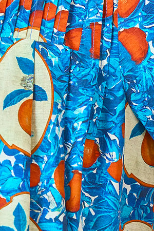 Close-up of pleated fabric with a vibrant orange grove print in blue and orange, perfect for the Girls' Youjin Dress by Hunter Bell NYC, featuring oranges, leaves, and oval frames.