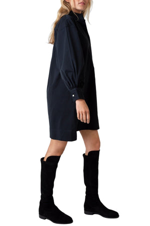 A person wearing an Ann Mashburn Anaya Popover Dress—featuring long puffed sleeves—and black knee-high boots.