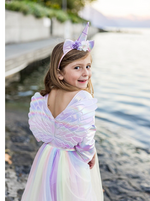 A young girl stands by the water, smiling back while wearing the Great Pretenders Alicorn Dress with Wings and Headband.