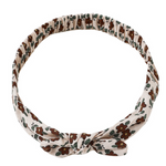 The Rylee & Cru Baby Bow Headband features a soft beige band with a brown and green floral pattern, knotted at the front. Its scrunchy elastic design ensures a comfortable fit for all-day wear.