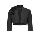 The Rose Bolero by Lela Rose is a black cropped jacket in a soft polyester-viscose blend, featuring three-quarter sleeves and two large fabric rose accents on the left shoulder. Imported for a modern, elegant look.