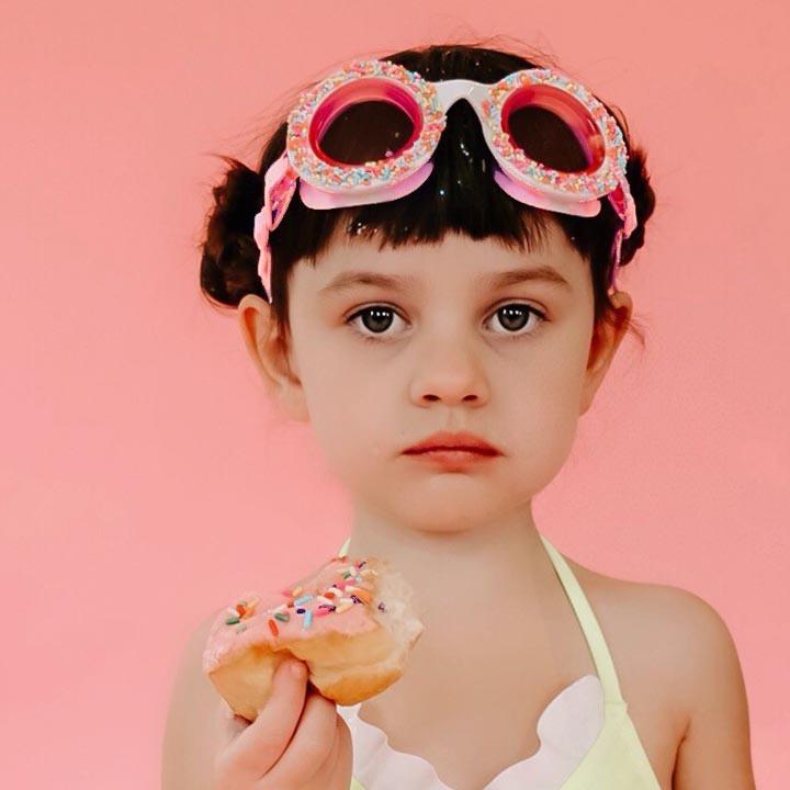 A young child wears Bling2O Do-Nuts 4 U Swim Goggles in Boston Creme Pink and a swimsuit, holding a sprinkled donut against a pink background with a neutral expression.