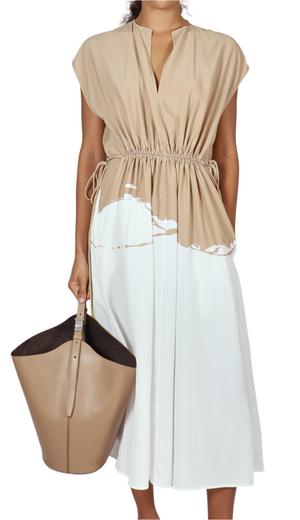 A person wears the Proenza Schouler Ezra Dress—a beige and white drawstring-waist style with a cinched silhouette—while holding a large beige tote bag.