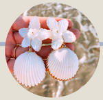 Close-up of a hand holding Collected By Farrell's La Mer Shell Earrings with white floral tops and shell-like pendants made from shimmering mother of pearl, against a blurred sea water background.