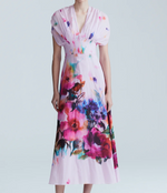 A woman wears the Lela Rose Watercolor Floral Cotton Voile Isabel Dress, featuring light pink cotton voile, short sleeves, a V-neck, and a watercolor floral print, as she stands against a plain light background.