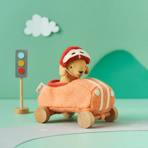 An Olli Ella Holdie Dog Go plush pup, sporting a red helmet, sits in an orange toy car with wooden wheels near a toy traffic light on a pastel green background.
