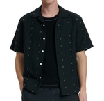A person wears the Avery Embroidered Camp Shirt by Rag & Bone in black cotton-linen over a black ribbed t-shirt, standing with hands in pockets against a plain background.