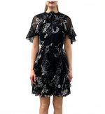 A woman wears the Jason Wu Collection SS Printed Chiffon Dress, a short black dress with white floral prints, ruffled short sleeves, and a high neckline, standing against a plain white background.
