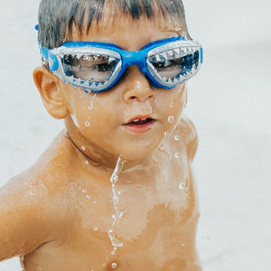 A young boy wears Bling2O's Jawsome Swim Goggles, featuring fun shark details and googly eyes, as he stands with water droplets on his face in a bright outdoor setting.