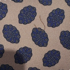 A beige Amoeba Tie by Robert Jensen featuring dark blue oval patterns and visible brown thread, evoking a hand-stitched silk design.