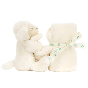The Jellycat Bashful Lamb Soother, crafted from recycled fibers, is nestled beside a rolled cream blanket tied with a ribbon.