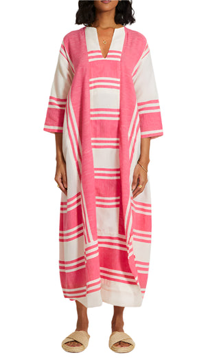 A model in the Lemlem Fana Caftan, featuring pink and white stripes and three-quarter sleeves, pairs it with open-toe woven slippers against a plain background for a versatile look.