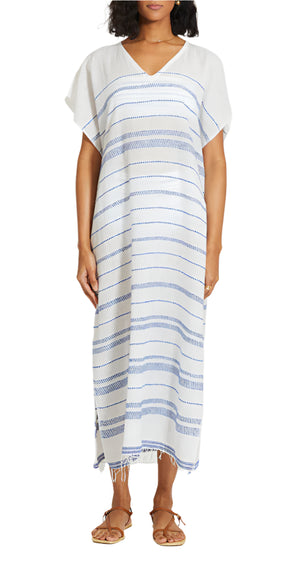 A woman wears the Lemlem Dalila Caftan—a hand-woven white dress with blue stripes, short sleeves, V-neck, and fringe hem—styled with brown sandals for a perfect beach cover-up.