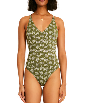 A woman showcases the Retiro One Piece swimsuit by Lemlem, featuring a stylish cross-back and V-neckline. The green jacquard design with a white palm tree pattern pops beautifully against the plain white background.
