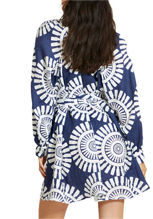 A woman wears the Lemlem Meaza Dress, featuring a blue and white pattern and long sleeves. Viewed from behind, her loose hair falls down her back as she stands with her hands at her sides.