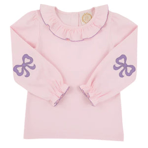 The Girls' Long Sleeve Ramona Ruffle Shirt by The Beaufort Bonnet Company features a light pink design with a ruffled collar, purple trim, and sweet bow accents—ideal for a stylish back-to-school outfit.