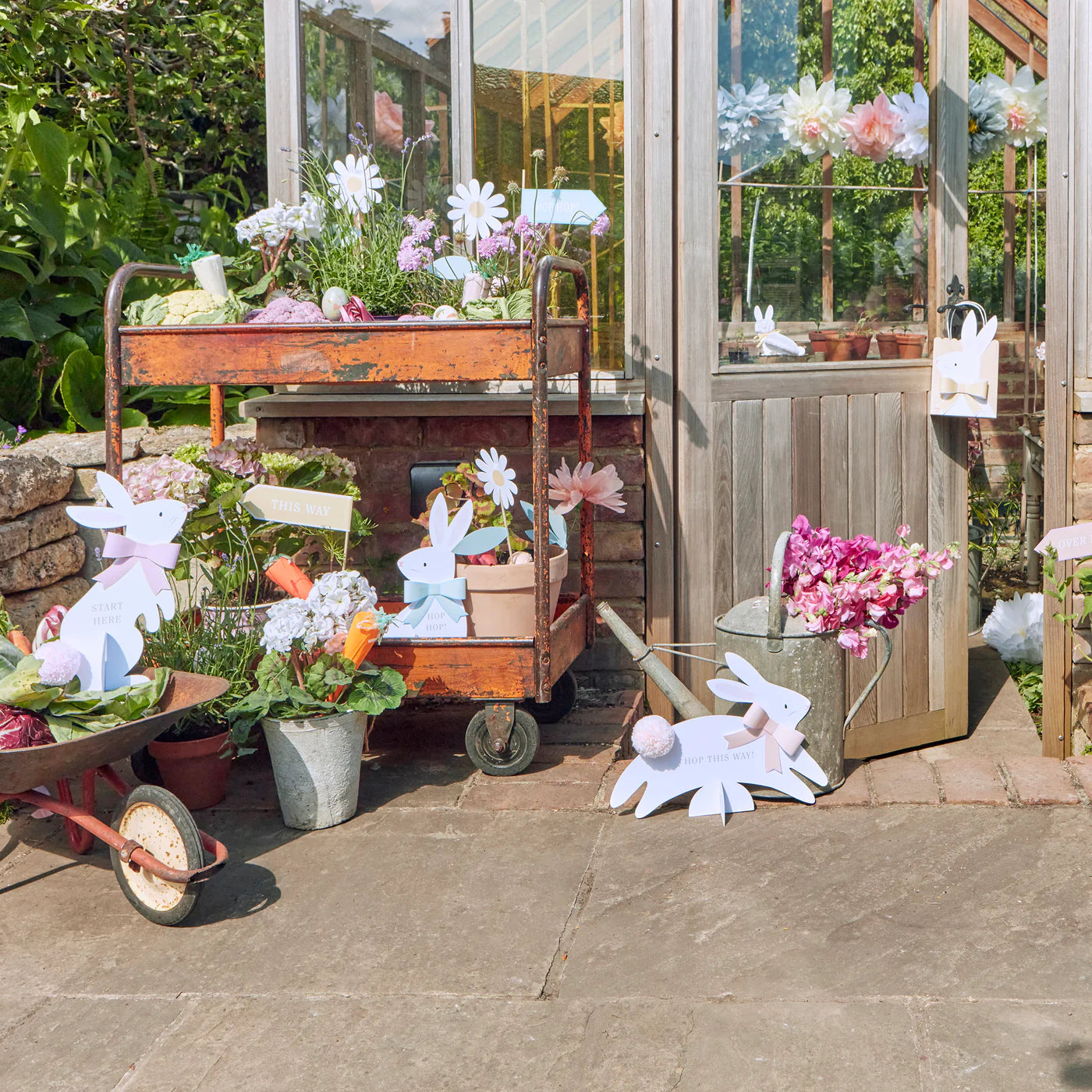 Outside a greenhouse, Meri Meri's Egg Hunt Kit adds charm with dapper bunny signs. Various plants and flowers are beautifully arranged on carts, baskets, and pots on a stone patio, creating an ideal setting for an Easter egg hunt.
