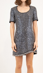 A woman wears the Simkhai Vivalie Mini Dress, a black and silver sequined checkered piece with short sleeves, a scoop neck, relaxed fit, and flattering silhouette, standing against a plain background.