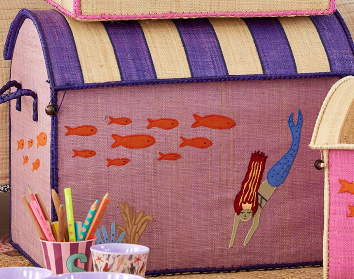 Rice's Raffia Toy Basket features a charming mermaid and fish design, crafted from Madagascar raffia in a purple and natural woven pattern.