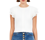 A woman wears the Ulla Johnson Giulia Top—a white, short-sleeve style with ruffle accents and subtle sheer knit—paired with high-waisted blue jeans with gold buttons.