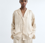 A person models Toteme’s Monogram Silk PJ Top, a loose-fitting beige blouse with a bold monogram pattern, paired with matching pants against a plain white background.