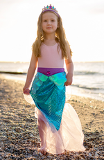 A child wearing Great Pretenders' Mermaid Glimmer Skirt and Tiara stands on a pebble beach by the sea during sunset.