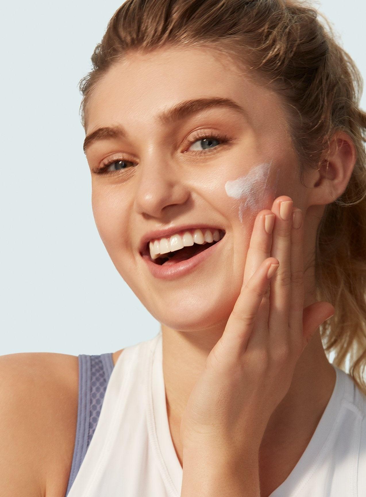 A smiling woman in a white tank top applies Supergoop Play Everyday Lotion with Sunflower Extract SPF 30, 2.4 oz., to her cheek with her right hand. Her light brown hair is pulled back.