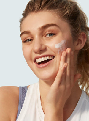 A smiling woman in a white tank top applies Supergoop Play Everyday Lotion with Sunflower Extract SPF 30, 2.4 oz., to her cheek with her right hand. Her light brown hair is pulled back.