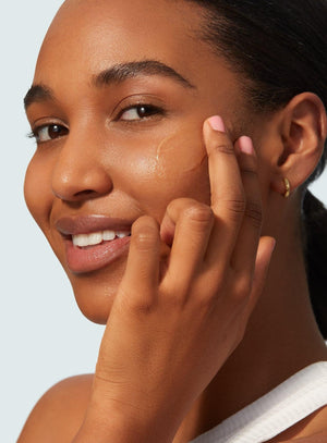 A woman in a white top smiles as she applies Supergoop Unseen Sunscreen SPF 40 to her cheek with her fingers against a plain background.