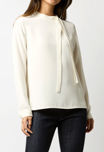 A person wearing the Ann Mashburn Pippa Top, a luxe crepe de chine tie-neck blouse, with dark pants, stands against a plain background.