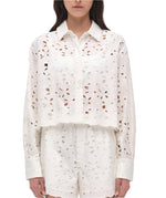A woman wears the Simkhai Penelope Shirt, a white, long-sleeve broderie anglaise top with delicate floral cutouts, paired with matching high-waisted shorts.