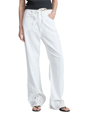 A person wears Rag & Bone's Ultra Featherweight Phoebe Drawstring Eyelet Jean with a drawstring waist and eyelet details near the hem, paired with black sandals and hands in pockets.