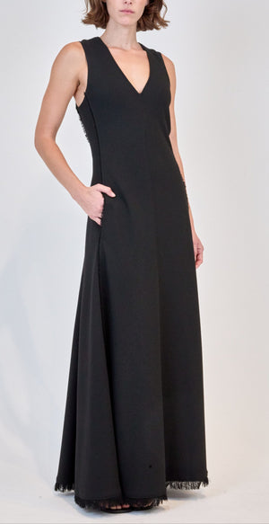 A woman wears the Simkhai Lowri Dress—a floor-length black gown with a V-neckline, sleeveless design, side pockets, and fluid silhouette—set against a plain light background.