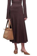 A person wearing the Simkhai Chiara Midi Skirt with a matching long-sleeved, ribbed brown top, holding a tan handbag and wearing tan heeled mules.