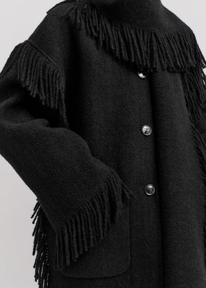 Close-up of Toteme's Fringed Scarf Jacket in black wool-blend bouclé, featuring fringe on the shoulders and sleeves, visible buttons, and a hand in the front pocket.