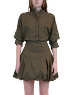 A person wearing the Effie Bubble Mini Dress by Simkhai—an olive green cotton poplin dress with a fitted waist, button-up top, and flared balloon skirt—stands against a white background.