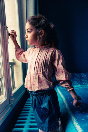 A young girl gazes out a window with one hand on the handle, wearing a light floral blouse and the Louis Louise Girls' Teacher Washed Denim Skirt featuring heart-shaped pockets and an elastic waistband.