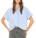 A person stands with one hand in their pocket, wearing the Drake Shirt from Xirena, a light blue button-front shirt made of crisp paper poplin, paired with dark gray pants.