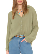 A woman wears a light olive green Xirena Scout Shirt, blue jeans, a brown belt, and layered necklaces with colorful charms.