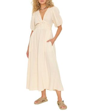 A woman wears the Xirena Lawsyn Dress—a cream-colored, short-sleeve cotton gauze midi with gathered waist and pockets—paired with flat sandals, standing against a white background.