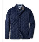 The Peter Millar Suffolk Coat, a navy blue water-resistant quilted jacket with a stand-up collar and snap buttons, is styled over a blue and white checkered shirt.