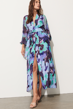 A person stands against a white wall in the Solana Dress by Figue, which features a long, floral pattern with a side slit. The silk chiffon fabric drapes elegantly, making it perfect for spring events.