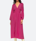 A woman wears the Sea NY Fabiola Dress, a magenta midi with long sleeves, deep v-neckline, gathered waist, and elegant puff sleeves.