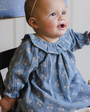 A baby wearing the Quincy Mae Baby Girls' and Girls' Long Sleeve Isla Dress sits on a chair, gazing upward with an open-mouthed expression.