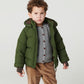 Young boy with curly hair in Copenhagen Colors Boys’ 2-in-One Puffer Jacket w/ Removable Sleeves, gray sweater, and brown corduroy pants, standing seriously against a plain background.