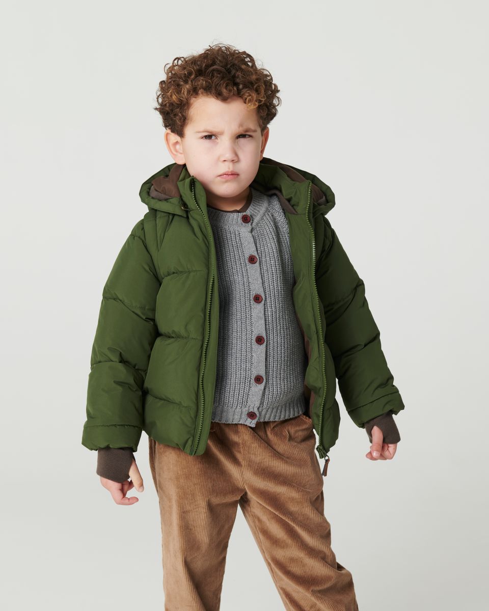 Young boy with curly hair in Copenhagen Colors Boys’ 2-in-One Puffer Jacket w/ Removable Sleeves, gray sweater, and brown corduroy pants, standing seriously against a plain background.