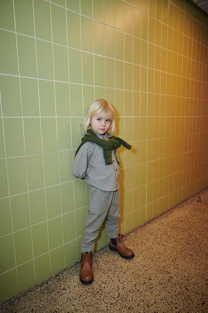 A young child with blonde hair wears the Baby Boys’ and Boys’ Checkered Classic Shirt by Copenhagen Colors under a light-grey suit, with a green sweater and brown boots.