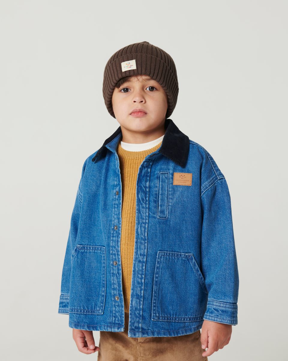 A young child wears the Copenhagen Colors Boys’ Denim Jacket with Corduroy Collar, a brown knit beanie, mustard sweater, and light brown pants against a plain background.