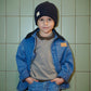 A young child stands against a tiled wall wearing the Copenhagen Colors Boys’ Denim Jacket with Corduroy Collar, a black knit hat, grey sweater, and blue jeans.