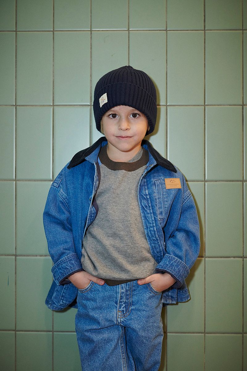 A young child stands against a tiled wall wearing the Copenhagen Colors Boys’ Denim Jacket with Corduroy Collar, a black knit hat, grey sweater, and blue jeans.
